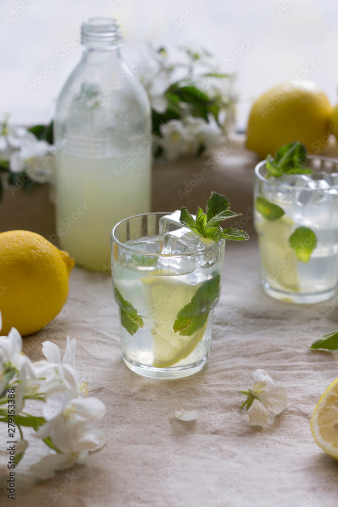 Fototapeta premium Bottle and glasses of cold lemon drink, ice, mint leaves, slices, white apple flowers on linen tablecloth. Close-up, copy space, vertical format
