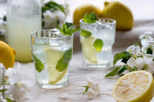 Bottle and glasses of cold lemon drink, ice, mint leaves, slices, white apple flowers on linen tablecloth. Close-up, copy space