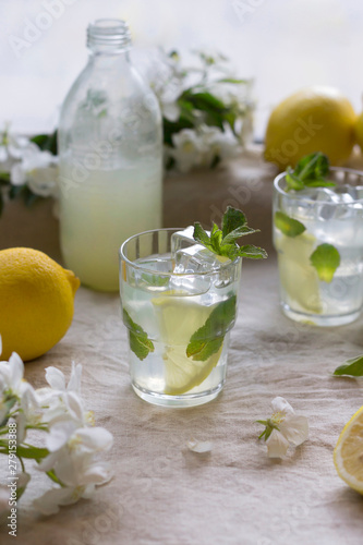 Bottle and glasses of cold lemon drink, ice, mint leaves, slices, white apple flowers on linen tablecloth. Close-up, copy space, vertical format