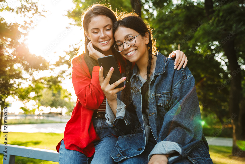 Image of two smiling girls using cellphone and hugging while sitting on railing in green park Image of two smiling girls using cellphone and hugging while sitting on railing in green park