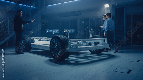 Automobile Design Engineers Talking while Working on Electric Car Chassis Prototype. In Automotive Innovation Facility Concept Vehicle Frame Includes Tires, Suspension, Engine and Battery © Gorodenkoff
