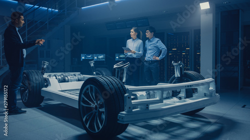 Automobile Design Engineers Talking while Working on Electric Car Chassis Prototype. In Automotive Innovation Facility Concept Vehicle Frame Includes Tires, Suspension, Engine and Battery © Gorodenkoff