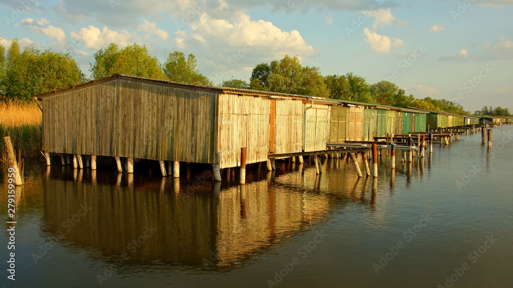 Fototapeta premium warmes Sonnenlicht erleuchtet viele Bootshäuser auf Stelzen in einem See in Mecklenburg