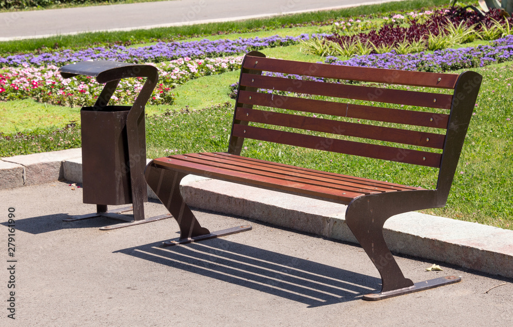 urn next to a street bench. Sidewalk with wooden bench and dumpster. Black metal wastebasket near the bench on the street urn next to a street bench. Sidewalk with wooden bench and dumpster. Black metal wastebasket near the bench on the street