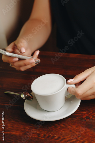 Cup of cappuccino on a wooden table next to the hand of the girl with the phone close up.