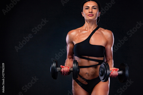 Strong muscular bodybuilder athletic woman pumping up muscles with dumbbells on black background. Workout bodybuilding concept.