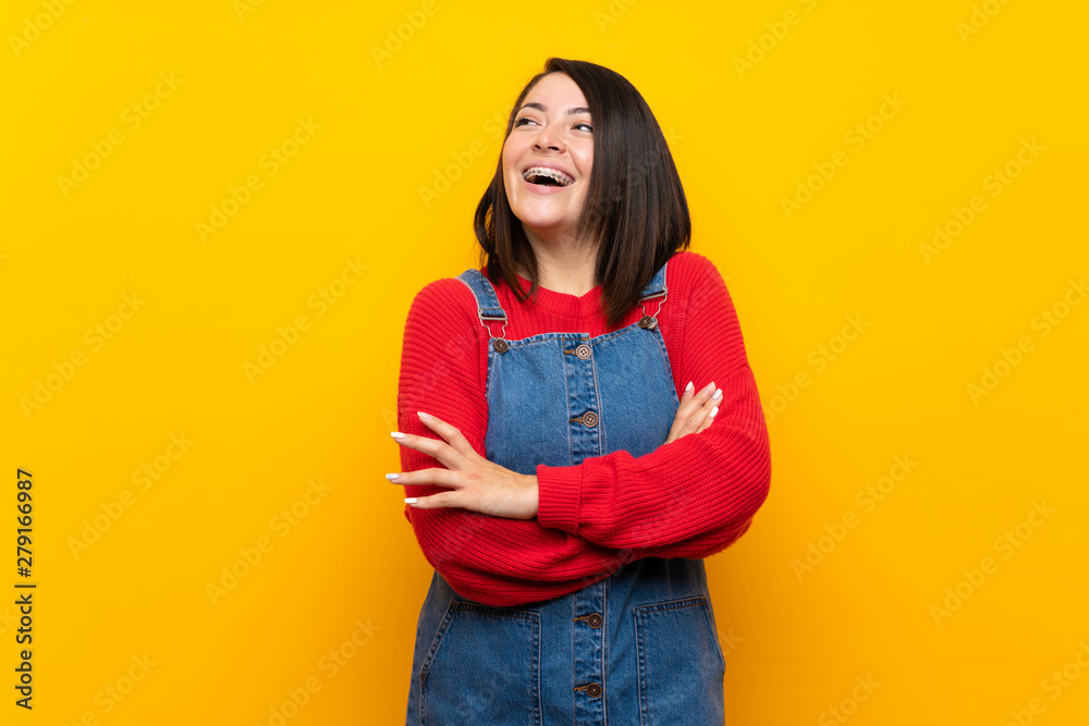 Young Mexican woman with overalls over yellow wall happy and smiling Young Mexican woman with overalls over yellow wall happy and smiling