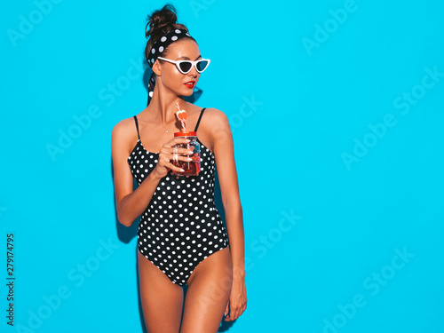 Young beautiful sexy smiling hipster woman in sunglasses.Girl in summer peas swimwear bathing suit.Posing near blue wall,drinking fresh cocktail smoozy drink