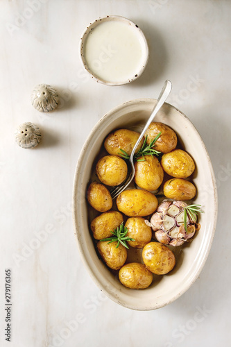 Young baked potatoes with garlic and rosemary in ceramic baking dish, served with cheese sauce, salt and pepper over white marble background. Flat lay, space