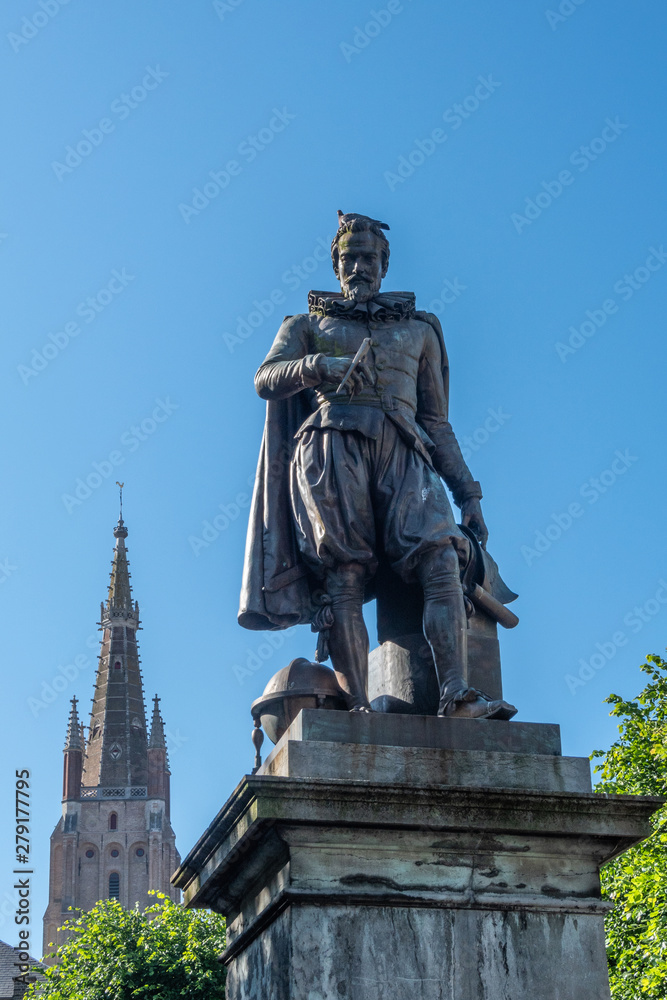 Bruges, Flanders, Belgium -  June 17, 2019: Simon Stevin statue with spire of OLV Cathedral in back under blue sky. Some green foliage. Pigeon on his head.