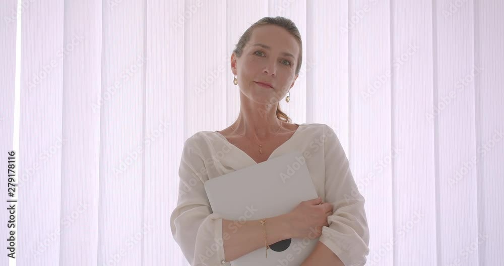 Closeup portrait of elderly caucasian brunette businesswoman holding a laptop looking at camera standing in a white apartment indoors