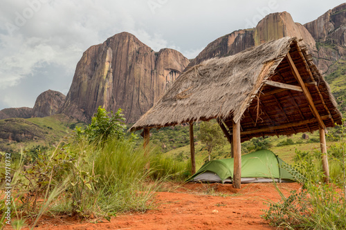 Small hiking tent pitched under an open thatched hut in the sacred forest, Tsaranoro valley, Madagascar. Famous rock formation mountain ridge on background  photo