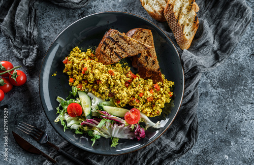 Vegan tofu scramble with vegetables, salad and toasted bread in plate over grey background. Healthy breakfast food, clean eating, vegan dieting, top view photo