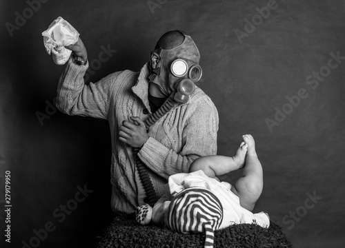 Young Dad cleaning his baby's dirty ass, changing the stinky diaper in a gas mask, fatherhood and humor..Father changes the baby's diapers on a changing table, on a dark, single track background.