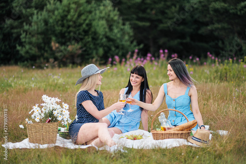 Three young women, blond, brunette and with dyed hair in blue dresses, and hats, sit on plaid and drink wine from glasses. Outdoor picnic on grass in forest. Delicious food in picnic basket.