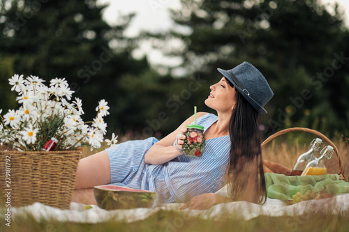 A young beautiful woman in a hat and dress is drinking lemonade from a can while sitting on a plaid on the green grass. Picnic basket, a bouquet of daisies, watermelon.