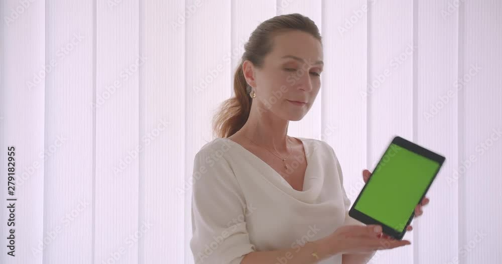 Closeup portrait of elderly caucasian brunette businesswoman using the tabletand showing green chroma key screen to camera standing in a white apartment indoors Closeup portrait of elderly caucasian brunette businesswoman using the tabletand showing green chroma key screen to camera standing in a white apartment indoors