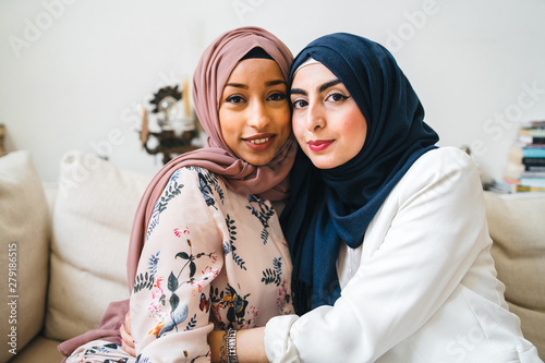 Portrait of two Muslim women with hijab on the sofa - Arab millennials embrace in an apartment