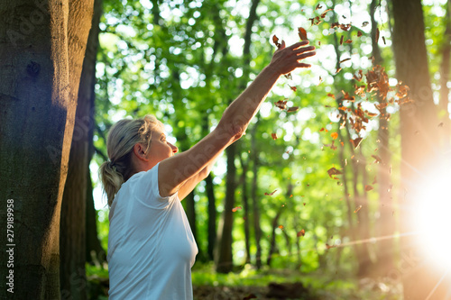 Experience the forest while bathing in the forest (Shinrin Yoku) with all her senses. A 50 year old blond woman plays with leaves. She throws it up. The evening sun is shining and she enjoys the at photo