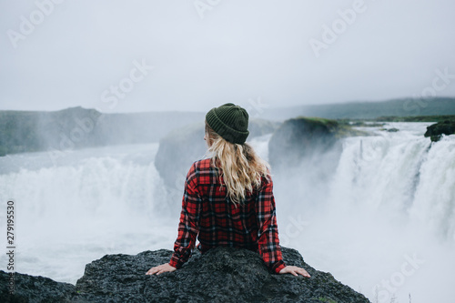 Canvas Print Beautiful young solo female adventurer or tourist rests on edge of cliff or mountain overlooking epic waterfall