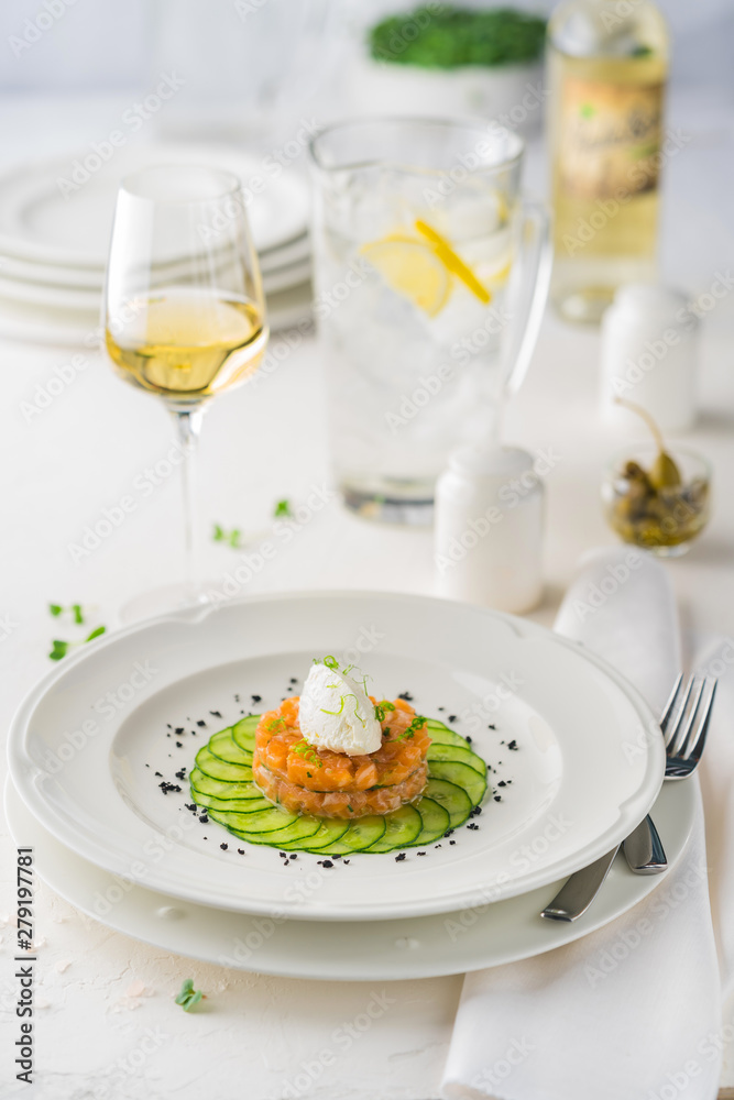 The salmon Tartar with cheese souffle on a white plate surrounded by Cutlery and white wine