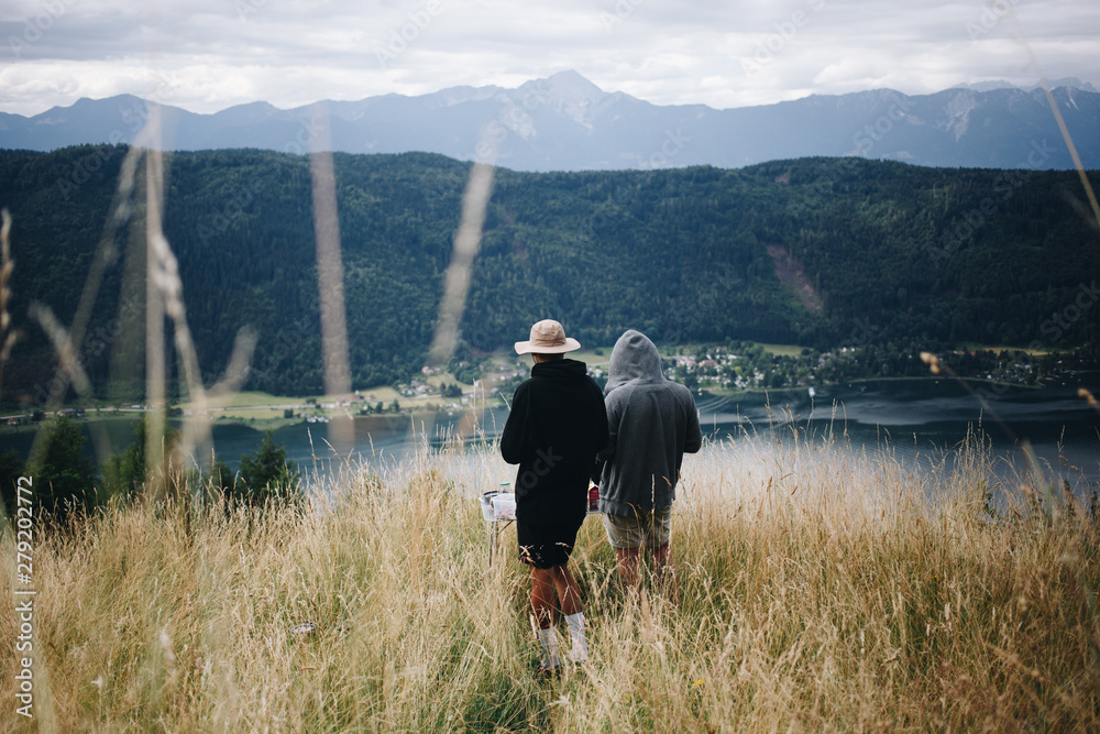 Two man in hiking or camping outdoor outfits stand in middle of alpine field, overlook valley with beautiful lake at bottom during summer vacation in national park