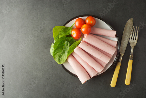 Thin slices of ham rolled on plate with fresh vegetables, dark background. Breakfast food, ingredient for sandwich. Flat lay food. photo