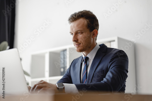 Calm young adult man in elegant suit working on his laptop