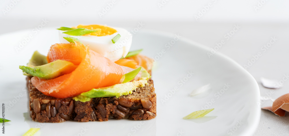 Healthy open sandwiches on multigrain wholegrain toast with avocado, salmon, eggs, herbs, sunflower seeds over white plate on concrete background with copy space. Healthy protein food. Wide banner. Healthy open sandwiches on multigrain wholegrain toast with avocado, salmon, eggs, herbs, sunflower seeds over white plate on concrete background with copy space. Healthy protein food. Wide banner.