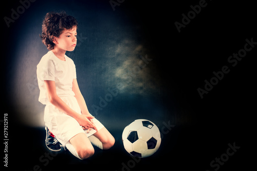 Sad and not happy little kid with football about lost football or soccer game. Dark background. Copy space. © kravik93