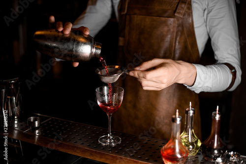 Professional bartender pouring a red alcoholic drink from the steel shaker through the sieve