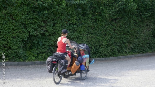 Trveling with bike and children. Young mother with two toddlers, tent and los of luggage rides on a load bicycle with electric motor on a country road.