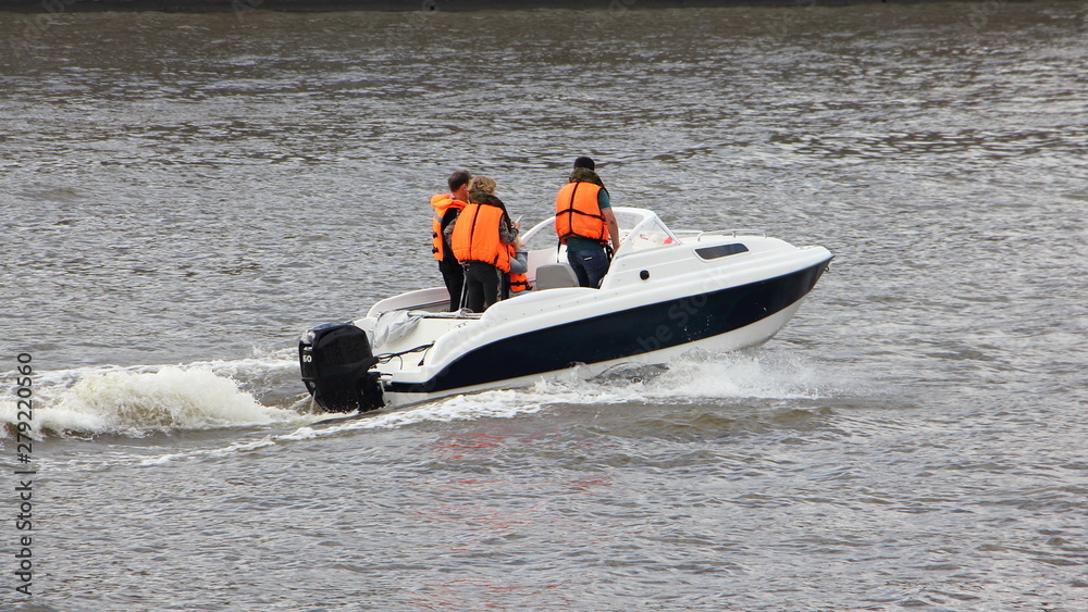 Fototapeta premium White plastic cabin motor boat with passengers in orange life jackets quickly floats on the river on a summer day