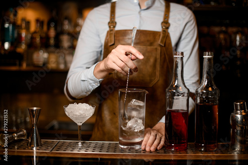 Professional male bartender stirring an alcoholic drink with ice in the measuring glass cup