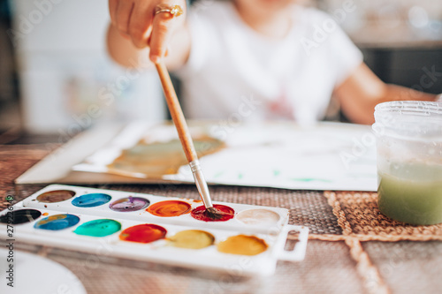 Beautiful blonde girl painting with paintbrush and water colors in the kitchen. Kid activities concept. Close up. Toned.