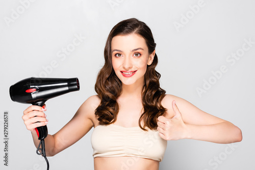 beautiful brunette woman with curls and makeup holding hairdryer and showing thumb up isolated on grey