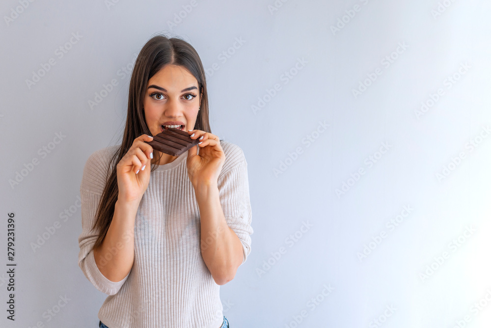 Portrait of a satisfied pretty girl biting chocolate bar and looking at camera isolated over white background. Smiling young lady standing over white background while holding chocolate Portrait of a satisfied pretty girl biting chocolate bar and looking at camera isolated over white background. Smiling young lady standing over white background while holding chocolate