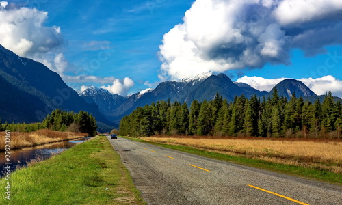 A country road of the Pitt River Valley runs through farm fields and forest to  Pitt Lake on the background of the snow-covered mountain range and a beautiful cloudy sky