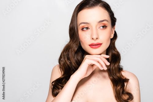 dreamy naked beautiful brunette woman with curls and makeup touching chin isolated on grey