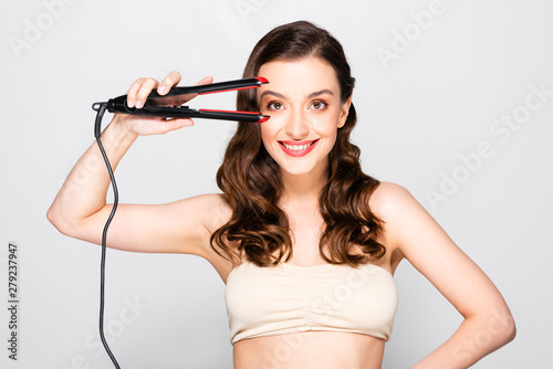 smiling beautiful brunette woman with curls and makeup holding straightening flat iron isolated on grey