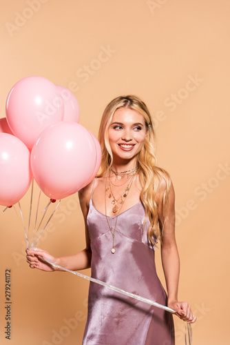 happy elegant blonde woman in violet satin dress and necklace holding pink balloons on beige
