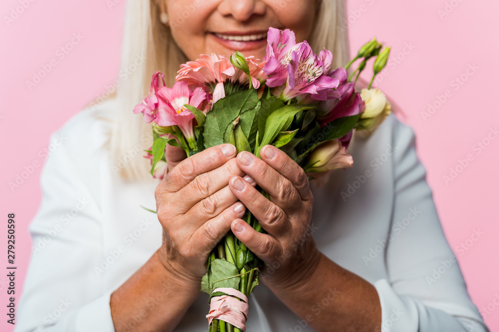 cropped view of positive senior woman holding bouquet of blooming flowers isolated on pink