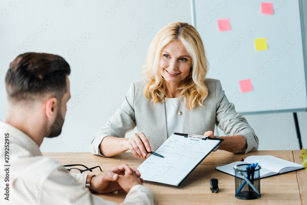 selective focus of happy recruiter holding pen near clipboard and looking at bearded man selective focus of happy recruiter holding pen near clipboard and looking at bearded man