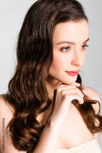 beautiful brunette woman with curls and makeup looking away with hand near chin isolated on grey