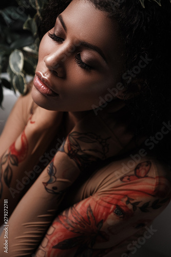 Glamour portrait of a metis young woman with perfect smooth glowing skin, full lips and fresh makeup on her face. Sensual and seductive african american female model is posing in the foliage