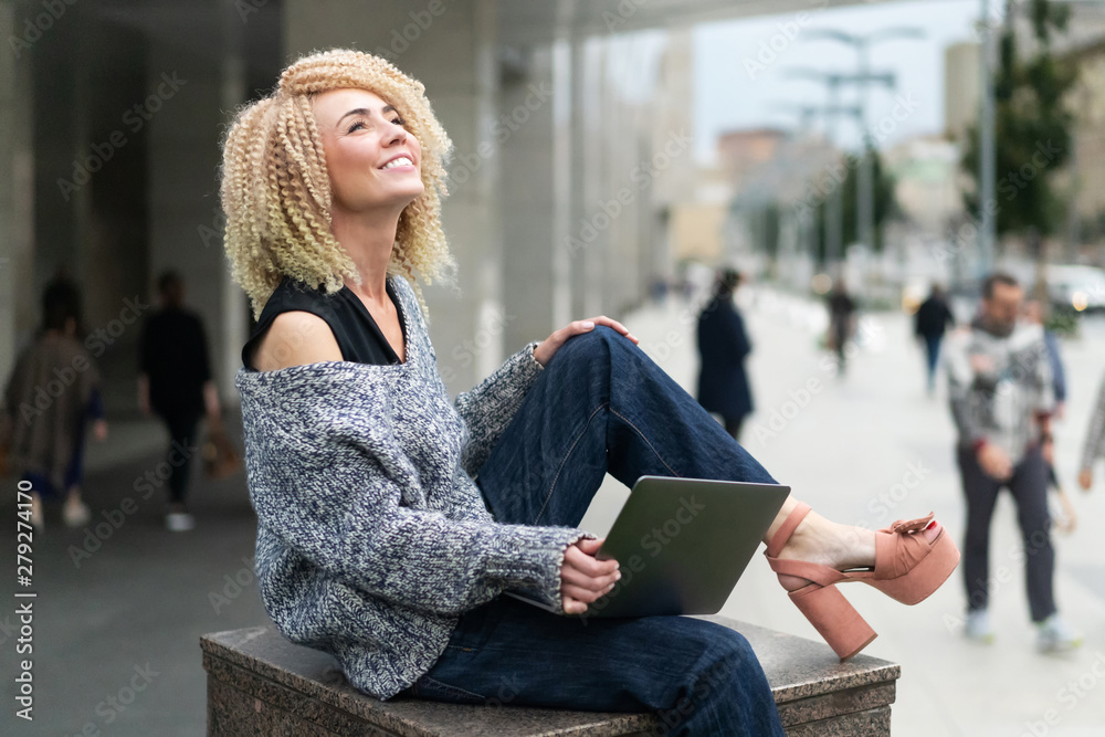 Obraz premium Young interesting hipster girl in sweater, jeans and hairstyle of afro curls sitting on parapet with laptop and working online on the remote. Freelancer. student learning