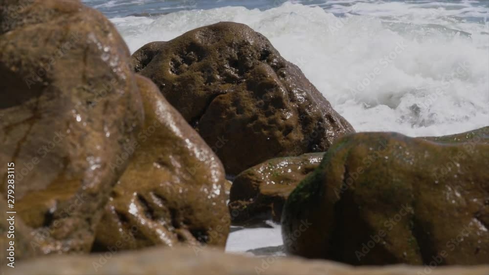 This slow motion video shows beach ocean waves crash over large brown rocks.