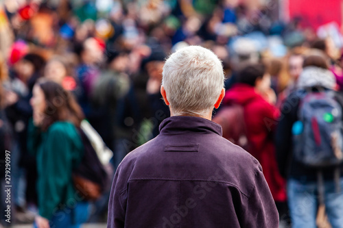 Ecological activists unite for climate. An older man is viewed from the rear with grey hair, contemplating a crowd of environmentalists marching in the city, people seen blurry in the background