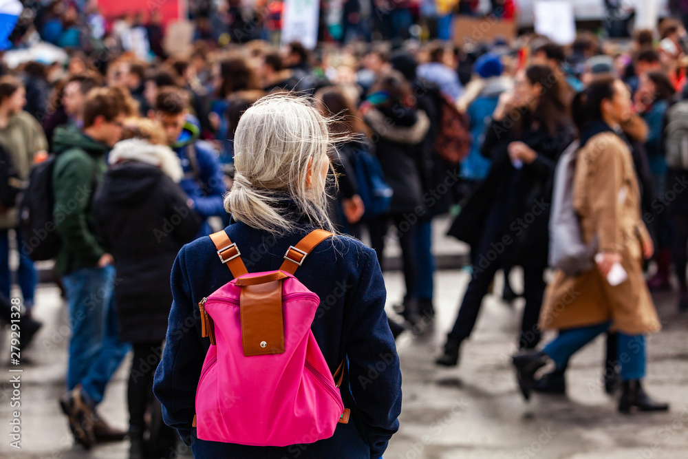 Person watches ecological protest. An older woman is viewed from behind, wearing a pink shoulder bag, watching an environmental demonstration on a street in Montreal, Canada