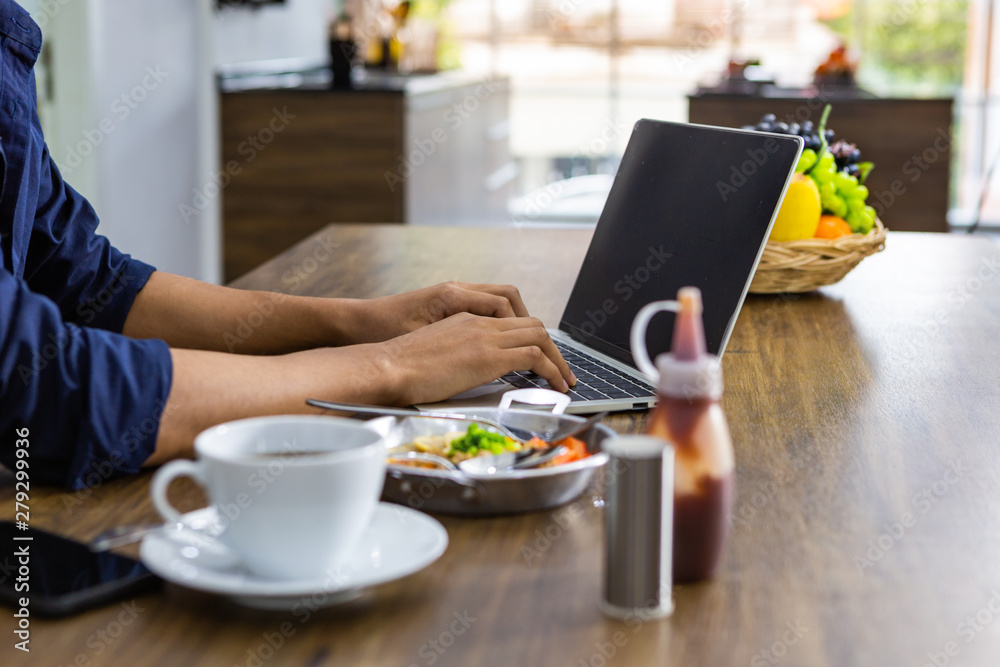Closeup of young asian man working laptop eating breakfast drinking coffee on wooden table in kitchen room Closeup of young asian man working laptop eating breakfast drinking coffee on wooden table in kitchen room